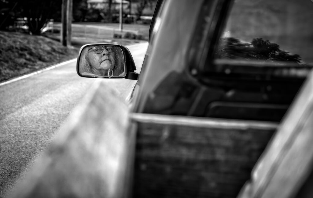 a black and white photo of a person driving a truck. the photo focuses on the side mirror and the woman is driving with conviction.