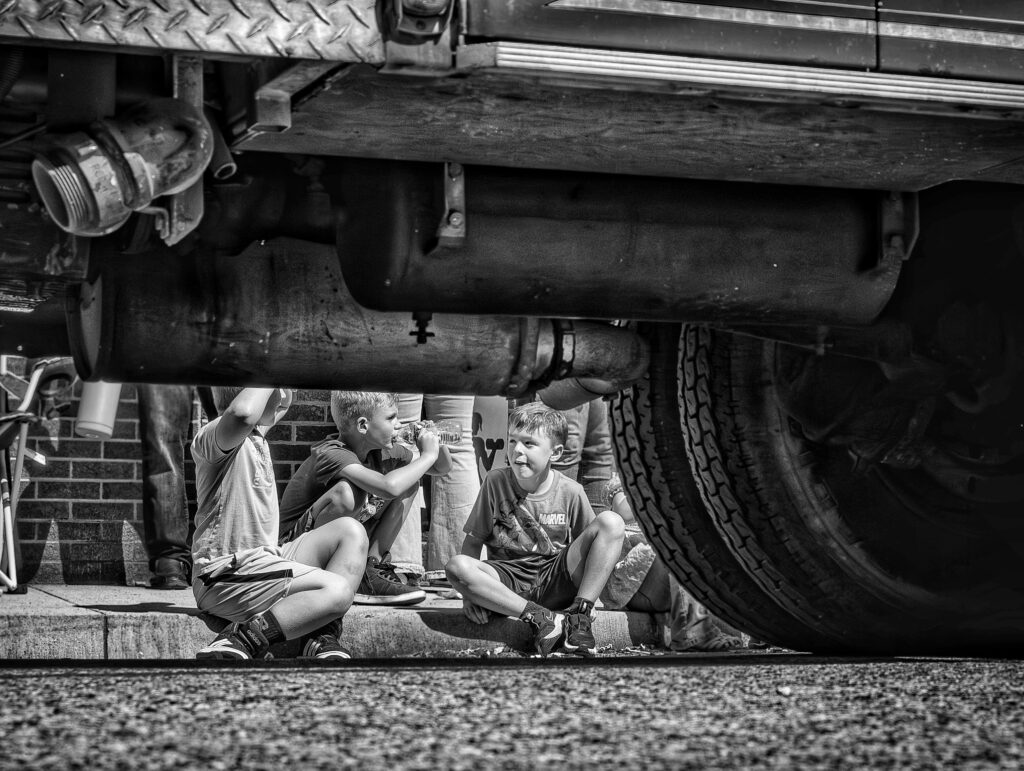 a black and white photo looks under a truck at three kids sitting on a curb