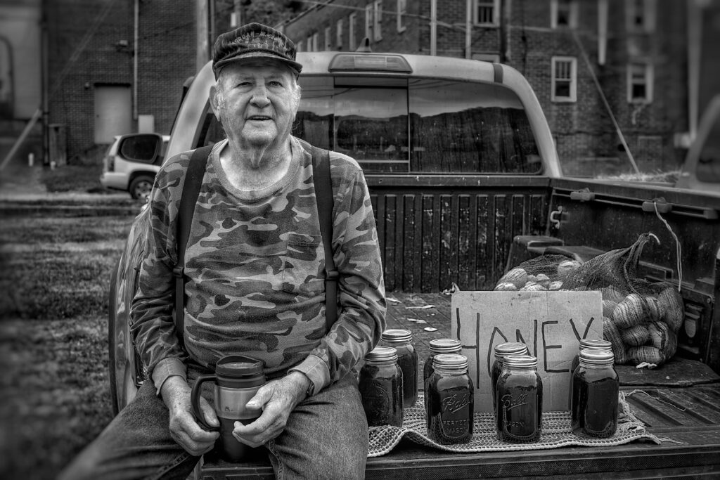 a black and white photo of a man selling honey our of the back of his truck. he's smiling and wears a camo shirt