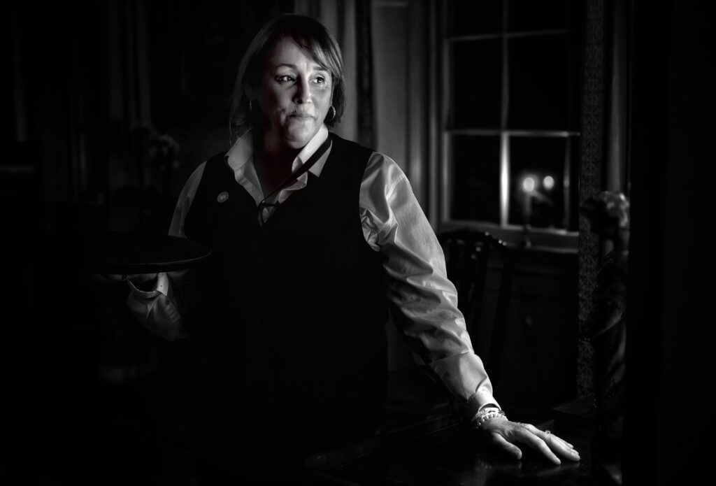 a black and white image of a person looking out a window. she wears a black vest over a white button down shirt. 