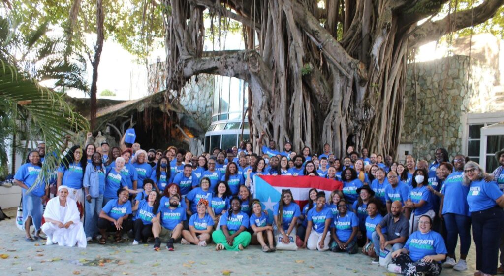 a multi-ethnic group of people are sitting on sand in front of a huge tree. Most of them are wearing a blue shirt that says "Gulf South 2 Appalachia" and they are holding the blue, red, and white flag of Borikén. 